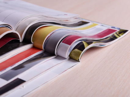 Kiosk und Lottoannahme Verschiedene geöffnete Zeitschriften die übereinander gestapelt sind und auf einem Holztisch liegen.