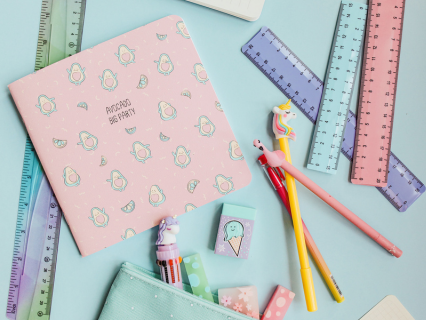 Schreibwaren Schreibwarensortiment. Lineal in Regenbogenfarben, ein rosa Notizheft mit kleinen Avocademännchen, verschiedene bunte Stifte mit kleinen Tieren als Radiergummis. Anschnitt eines geöffneten Mäppchens woraus Schreibwaren herauskommen.