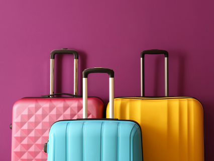 Taschen & Koffer Sortiment Drei Hartschalenkoffer in rosa, blau und gelb, stehend mit ausgezogenem Griff. Bildhintergrund burgunder.